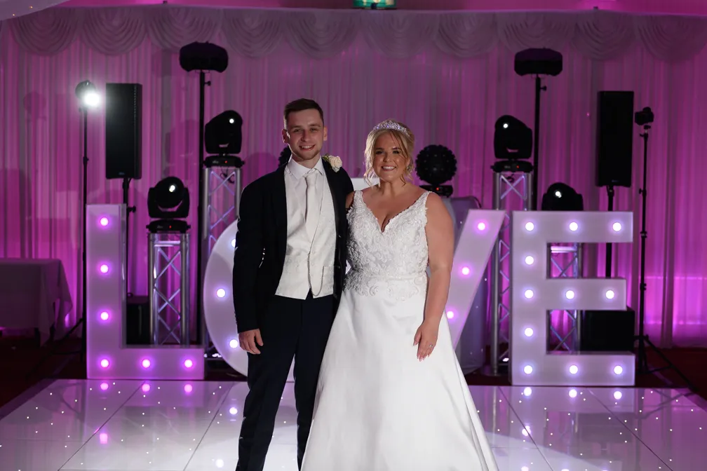 Bride in white gown and groom in dark suit standing together in front of large illuminated LOVE letters with purple lighting at wedding reception.