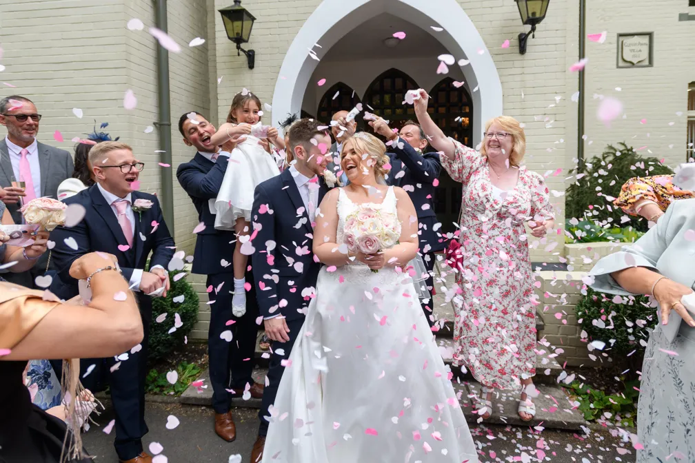 Bride and groom smiling as guests throw pink and white flower petals outside a building with an arched entrance.