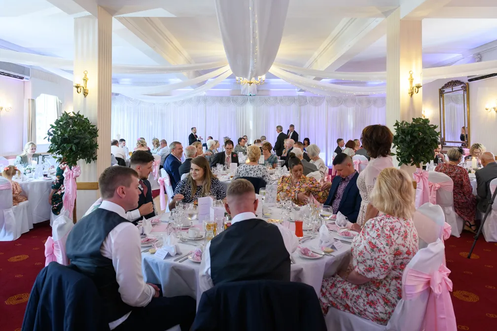 Wedding reception with guests seated at round tables covered in white cloths and pink bows on the chairs in a decorated hall.