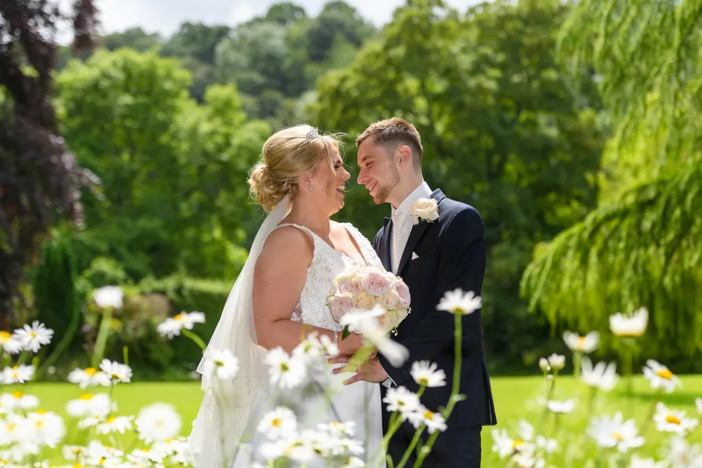 Bride and groom smiling and holding hands in a garden with white daisies and green trees.
