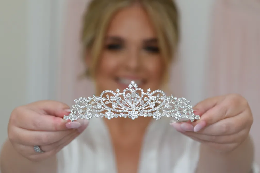 Woman holding a sparkling silver tiara with intricate floral designs, smiling in the background.
