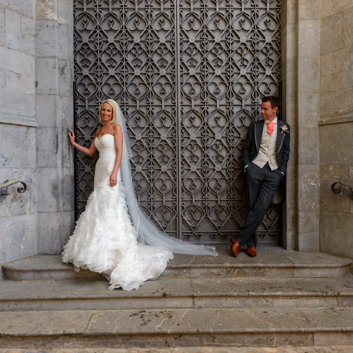 Bride in a strapless white ruffled wedding gown with a long veil and groom in a suit with coral tie standing on stone steps in front of ornate metal doors.
