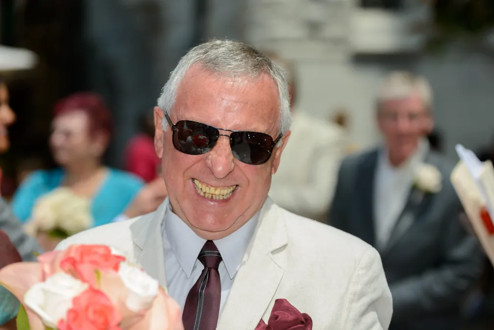 Smiling older man wearing sunglasses, a light-colored suit, and burgundy tie holding a bouquet of pink and white roses at an outdoor event.