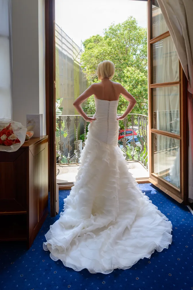Bride in a strapless white wedding dress with a ruffled train standing in a doorway facing a balcony with greenery outside.