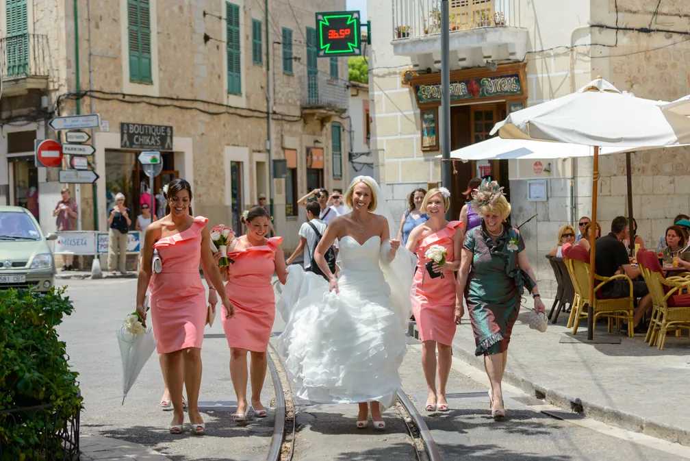 Bride in white wedding dress walking on street with bridesmaids in pink dresses and a woman in a teal dress, near outdoor café.