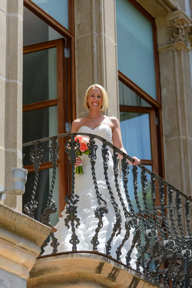 Smiling bride in a strapless white wedding gown holding a bouquet of orange and white flowers, standing on a stone balcony with ornate black iron railing and large windows behind her.