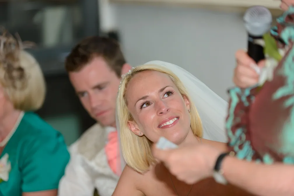 Bride smiling and listening attentively during a wedding speech, with a groom and a guest in the background.