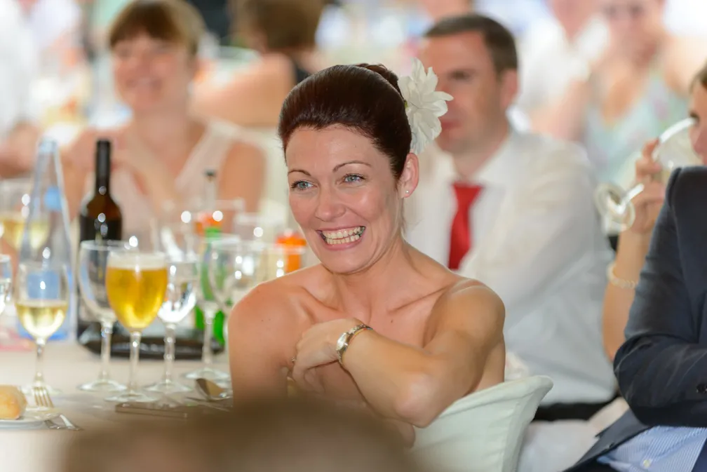 Smiling woman with flower in hair seated at a table during a social event with drinks and people in the background.