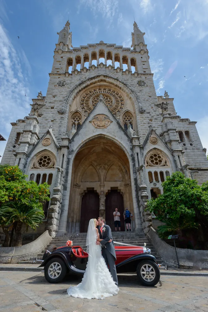 Bride and groom kissing in front of a large stone church with a vintage red and black car parked beside them.