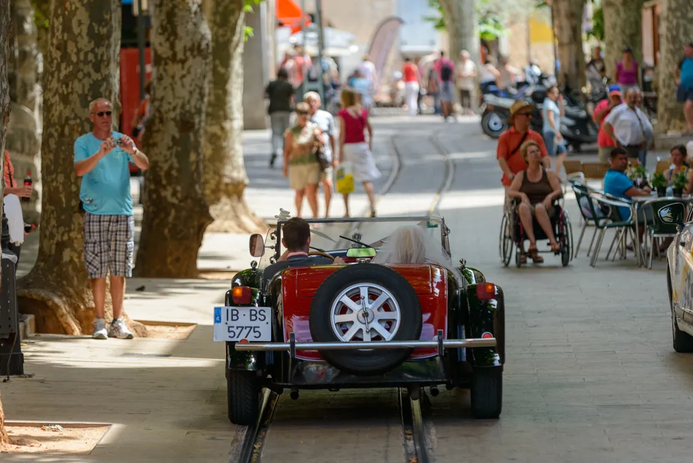 Rear view of a vintage convertible car driving on a tree-lined street with pedestrians and outdoor café seating.