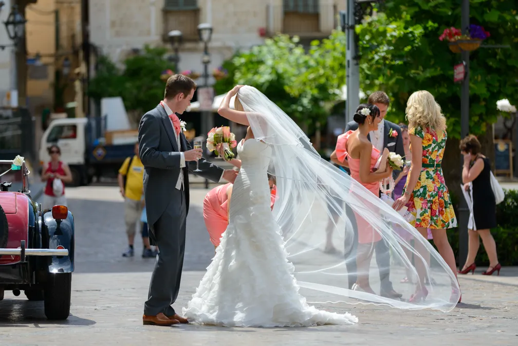 Bride in a white gown with a flowing veil and groom in a suit standing beside a vintage car, with wedding guests chatting in the background.