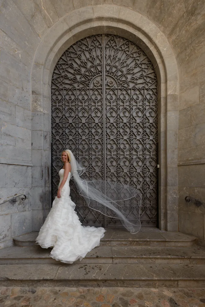 Bride in a strapless white wedding gown with a long veil standing on stone steps in front of an ornate large metal arched door.