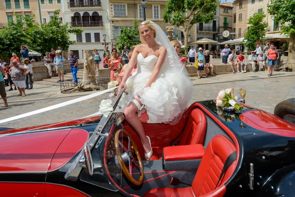 Bride in a white wedding dress sitting on the door of a red and black vintage convertible car in a town square with onlookers.