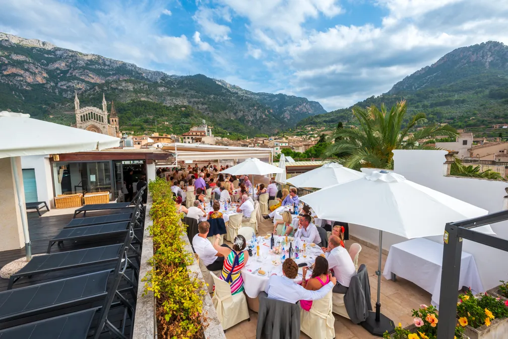 Outdoor rooftop dining area with guests seated at tables under white umbrellas, surrounded by mountains and buildings.