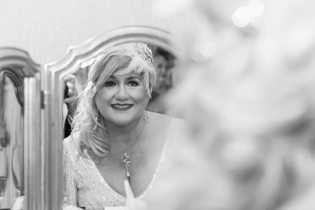 Smiling woman in a sequined dress with a feathered headpiece looking into an ornate mirror.