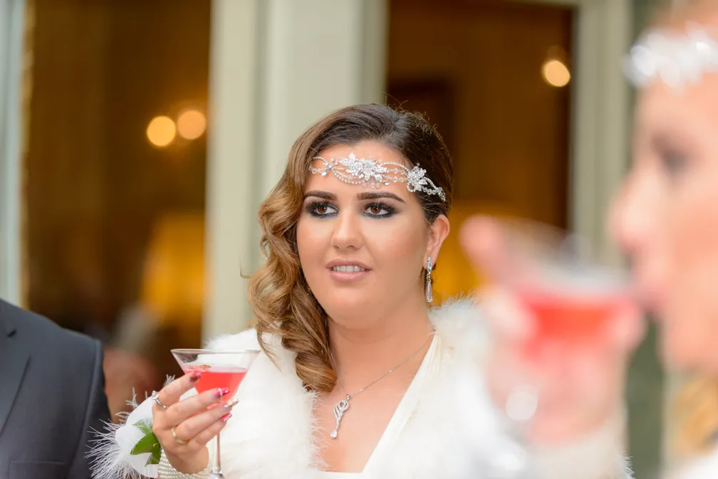 Woman with curled brown hair wearing a jeweled headpiece and white fur, holding a cocktail glass with a pink drink.