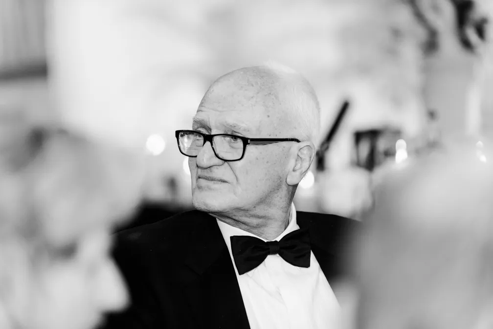 Elderly man with glasses wearing a tuxedo and bow tie looking to his right in a blurred indoor setting.