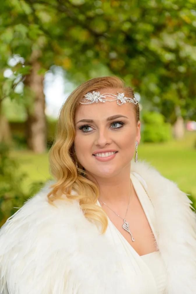 Smiling woman with wavy blonde hair wearing a white fur shawl, a jeweled headpiece, necklace, and earrings, posing outdoors with green foliage in the background.