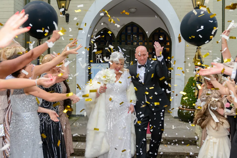 Newlywed couple walking out of a building while guests throw gold and white confetti during a wedding celebration.