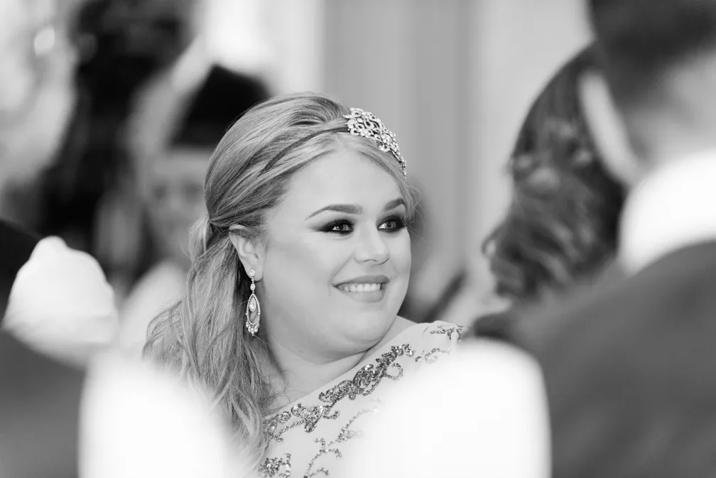 Smiling woman with styled hair, jeweled hair accessory, and earrings, looking to the side at an indoor event.