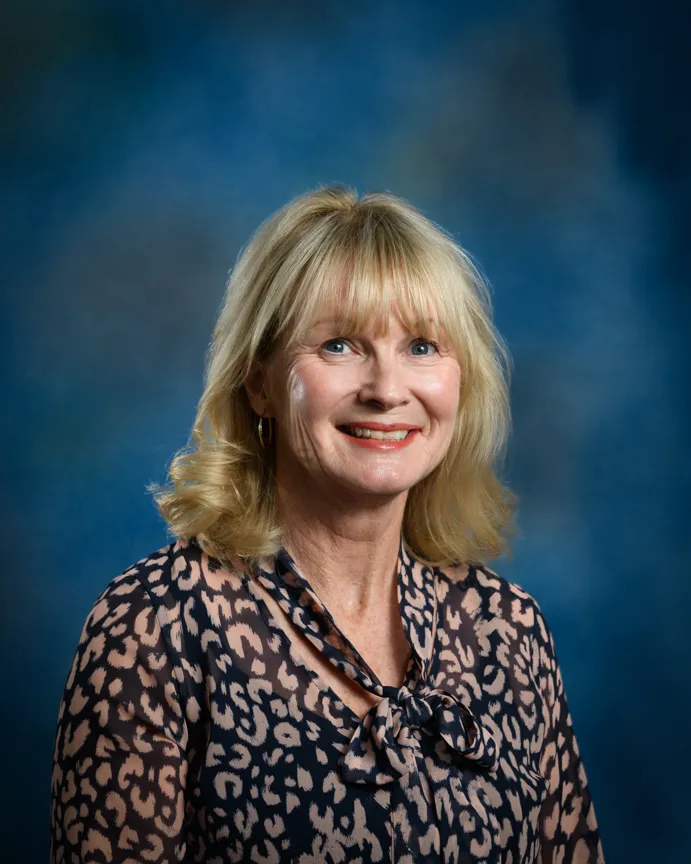 Smiling woman with blond hair wearing a patterned blouse with a bow tie collar against a blue studio background.
