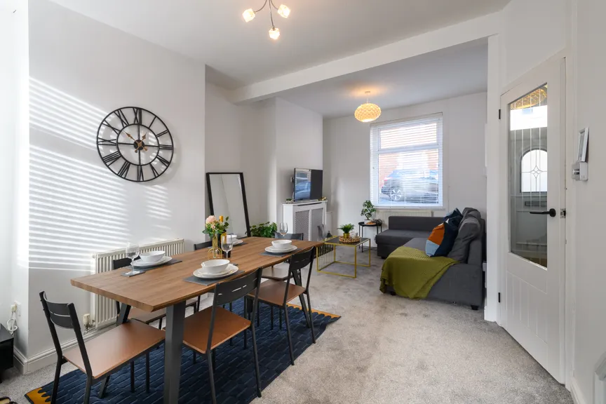 Modern dining and living area with wooden table set for four, grey couch with colorful pillows, a large wall clock, and window letting in natural light.
