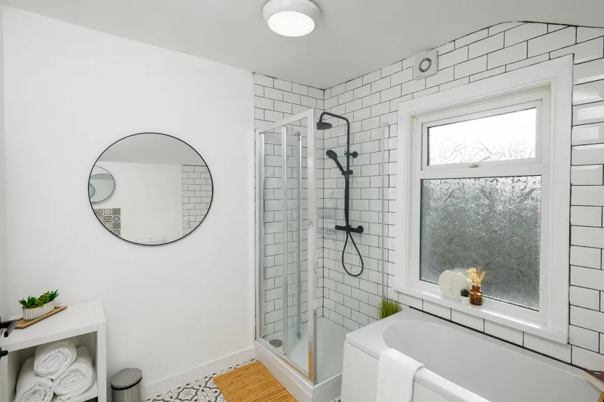 Modern bathroom with white subway tiles, glass shower enclosure, white bathtub with towel, round mirror, and small shelf with rolled towels and plants.