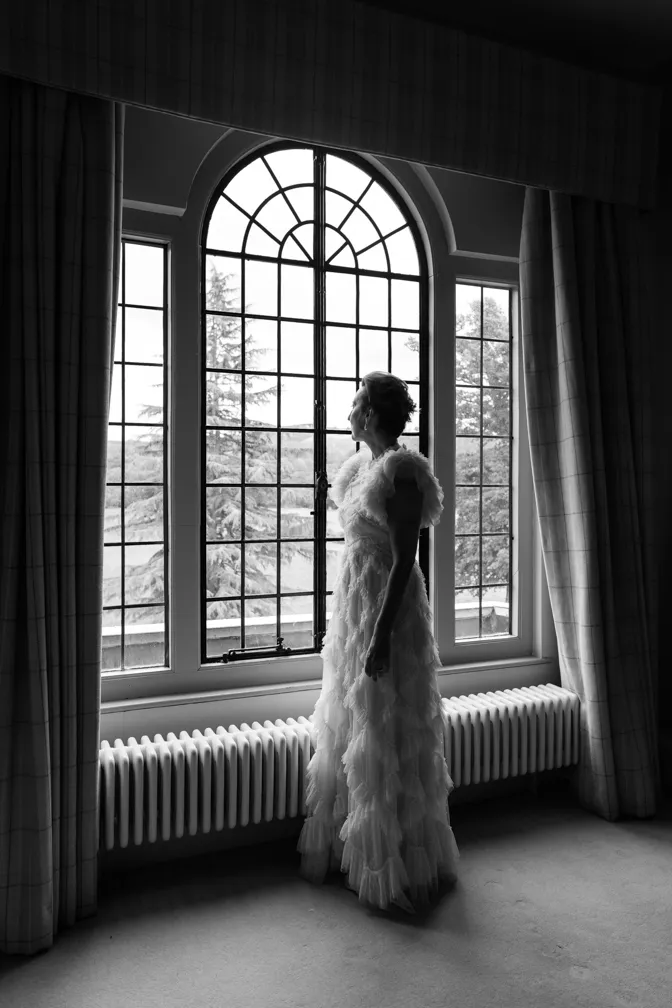Woman in a ruffled gown standing by a large window looking outside, with trees visible through the panes.