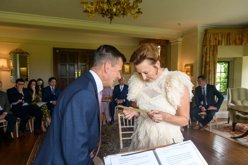 Bride in a white ruffled dress and groom in a blue suit smiling and reading a document together during an indoor wedding ceremony with seated guests in the background.