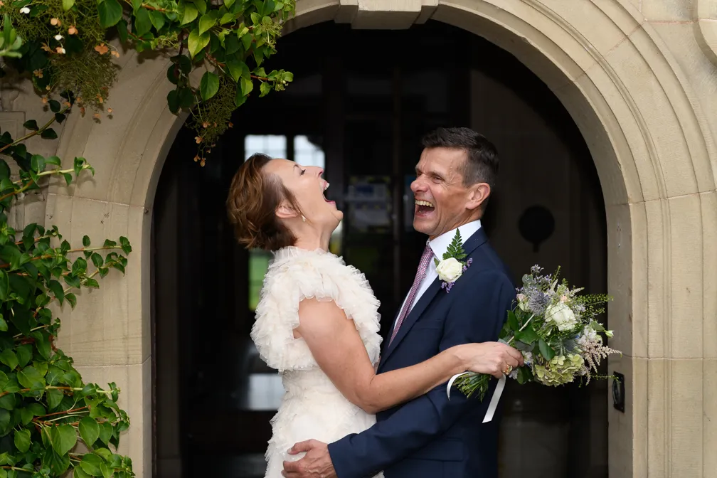 Bride and groom laughing and embracing under a stone archway adorned with greenery, bride holding a bouquet.