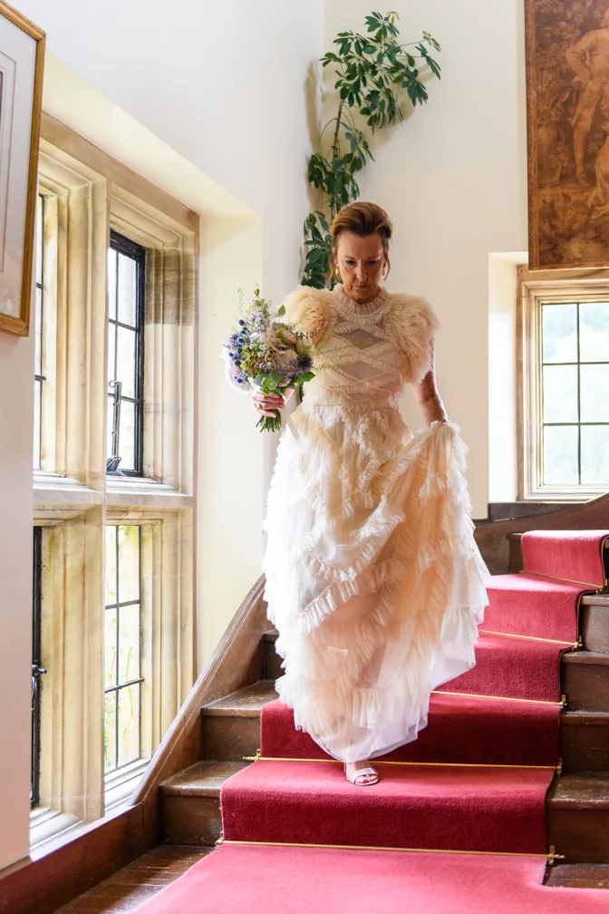 Woman in an elegant, layered cream dress holding a bouquet walking down red-carpeted stairs in a bright room with large windows.