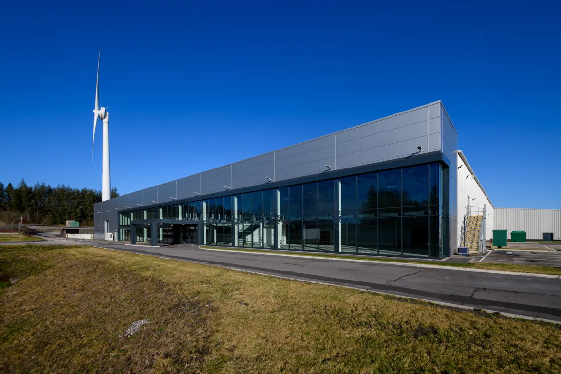 Modern industrial building with large glass windows next to a tall white wind turbine under a clear blue sky.