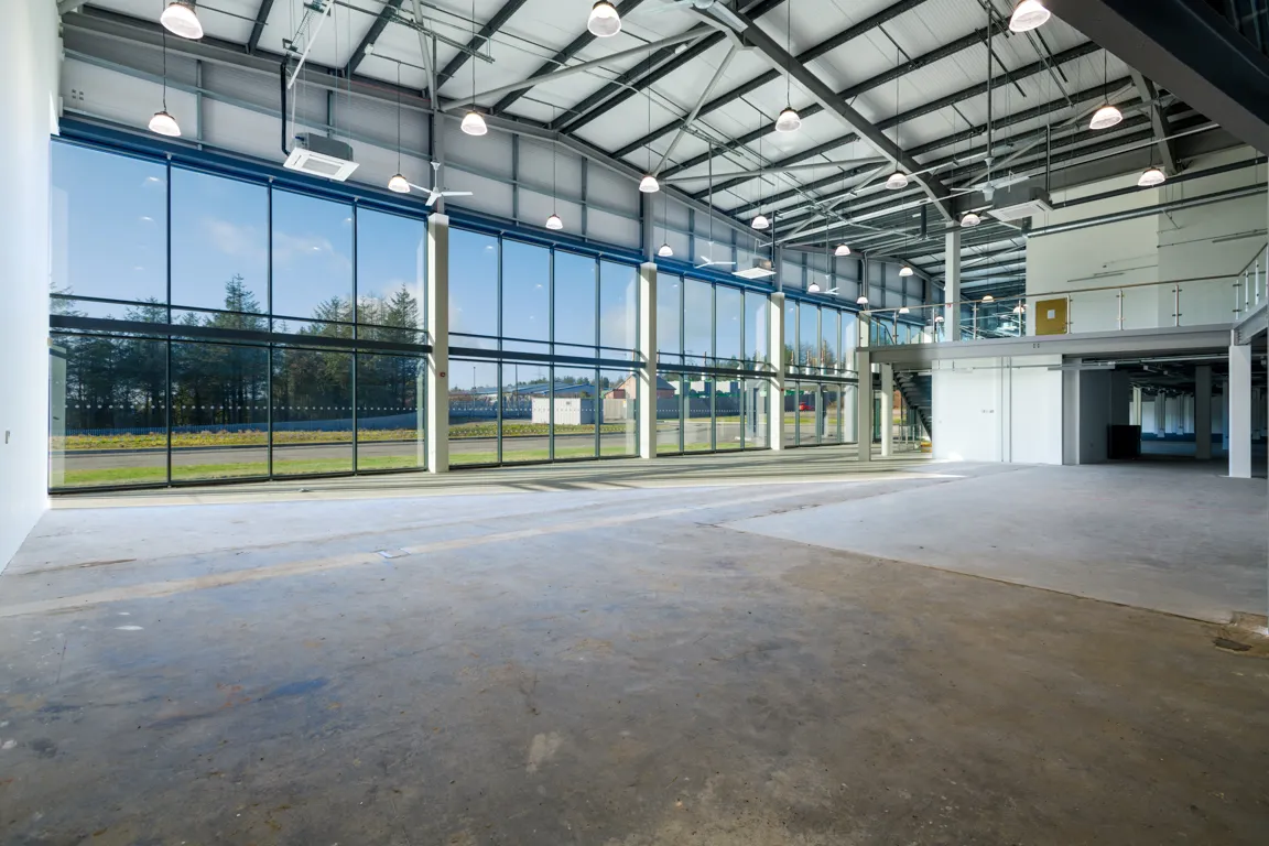 Spacious empty industrial warehouse with large glass windows and a mezzanine level with staircase.