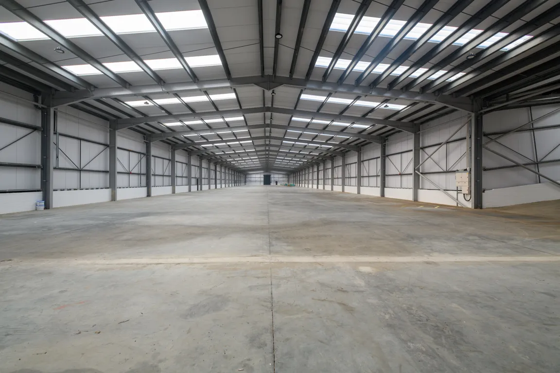 Spacious empty industrial warehouse with concrete floor and steel beams supporting a ceiling with skylights.