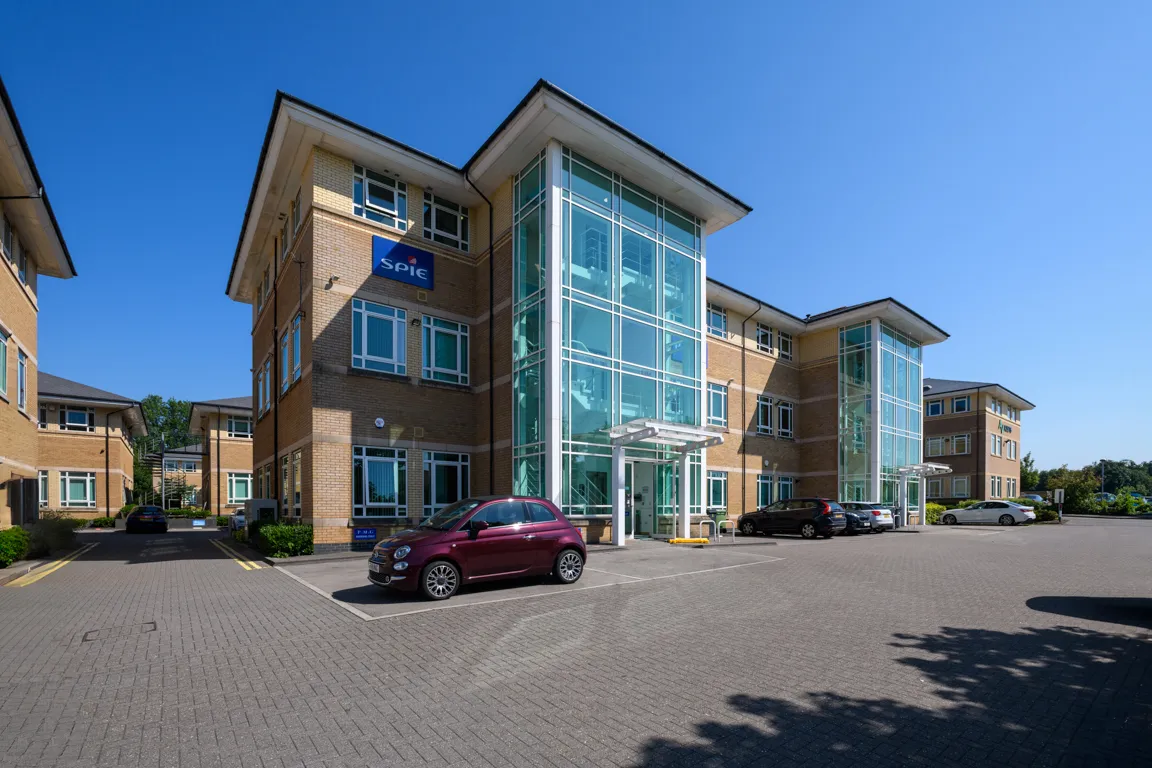 Modern office building with large glass stairwells, brick exterior, and several parked cars under clear blue sky.