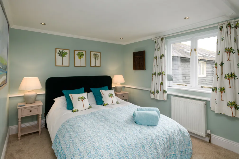 Cozy bedroom with light blue walls, a double bed with palm tree patterned pillows and curtains, two bedside lamps, and a window showing an outside building.