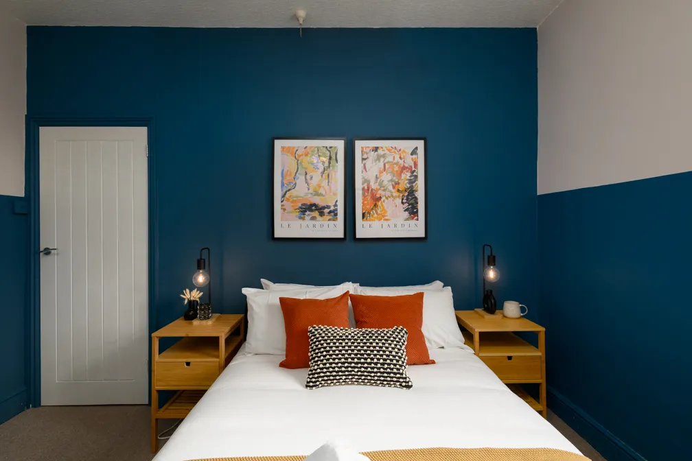 Bedroom with a white bed centered against a blue accent wall, featuring two orange pillows, a patterned black and white pillow, wooden nightstands with modern black lamps, and two abstract colorful framed artworks above the bed.