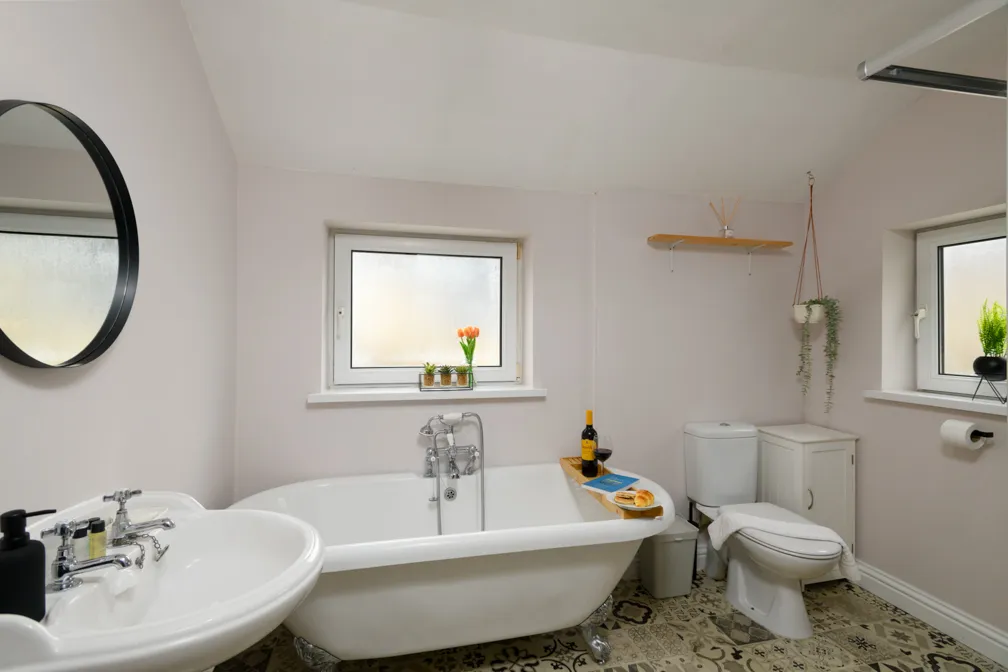White bathroom with a clawfoot bathtub, sink, toilet, patterned floor tiles, two frosted windows, and decorative plants.