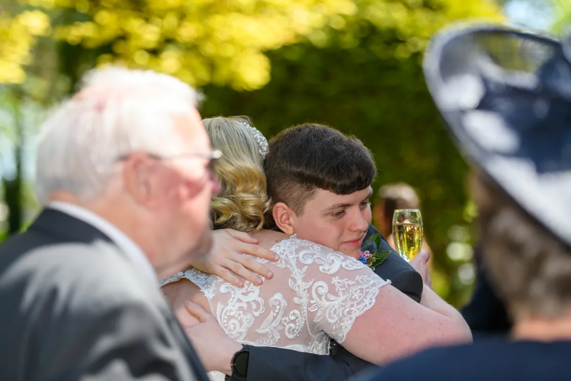 Young man in formal wear hugging a woman in a white lace dress, holding a glass of champagne, outdoors with blurred people in background.