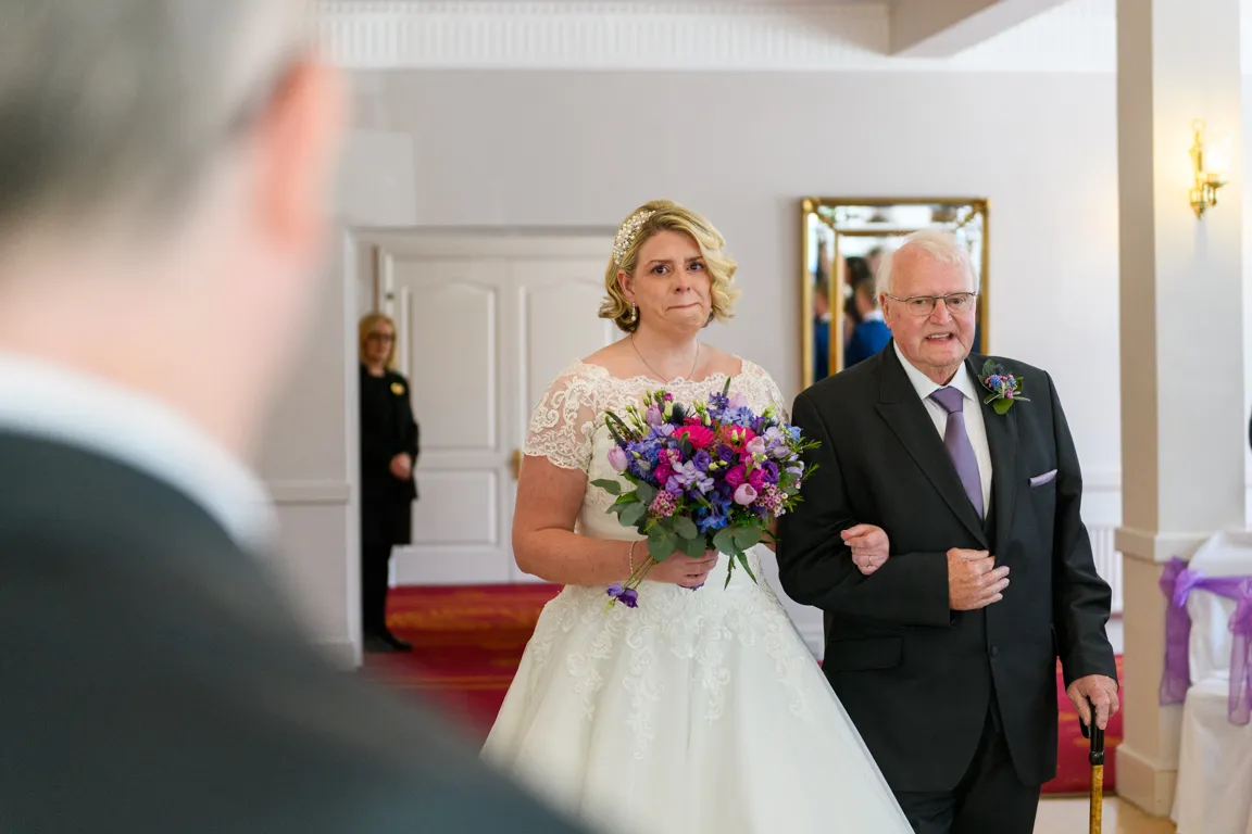 Bride in white wedding dress holding a bouquet of purple and pink flowers, walking arm-in-arm with an elderly man in a black suit.