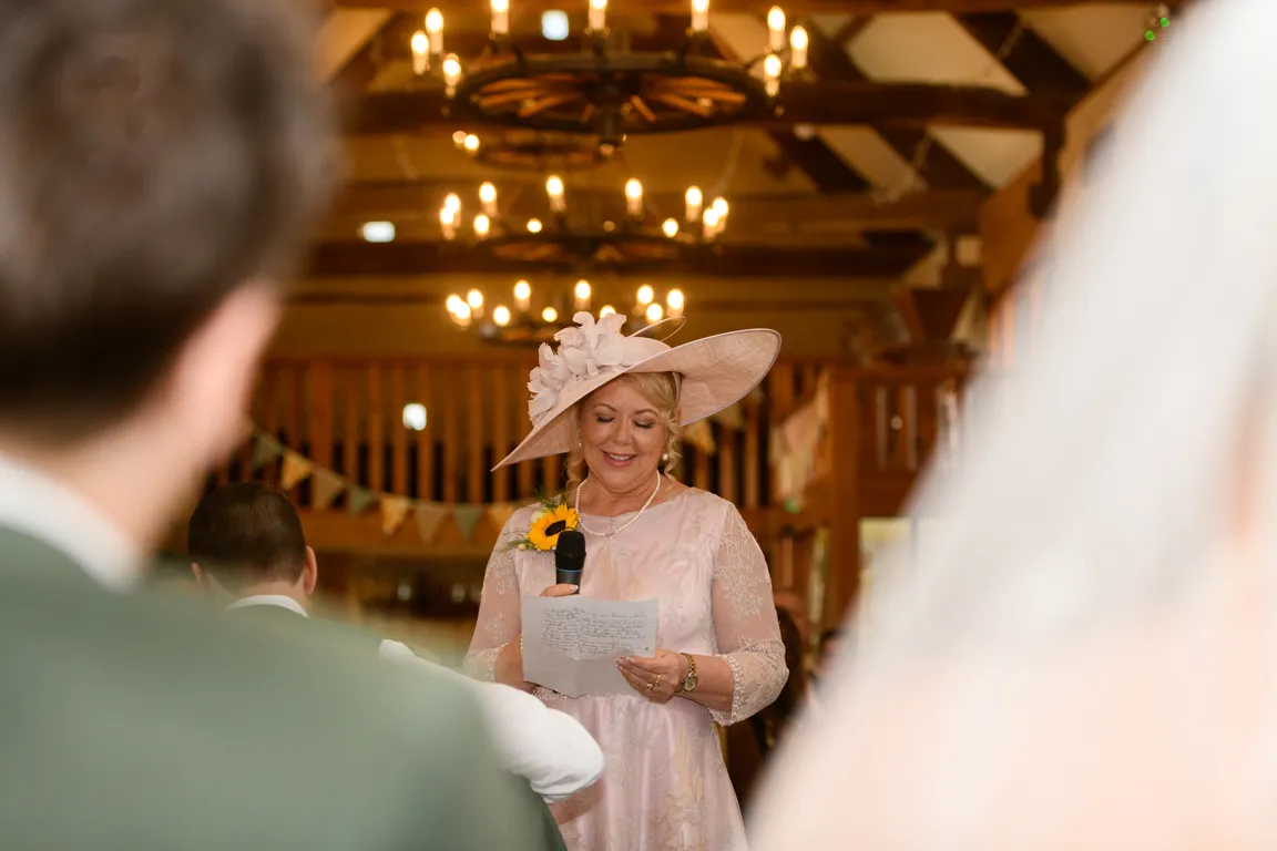 Woman in a pink dress and large hat with a sunflower corsage giving a speech with a microphone at a wedding reception.