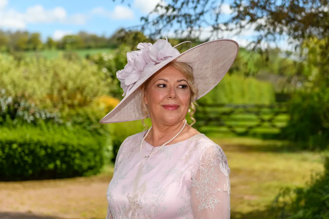 Smiling woman wearing a pale pink floral hat, pearl necklace, and lace dress standing outdoors with greenery in the background.
