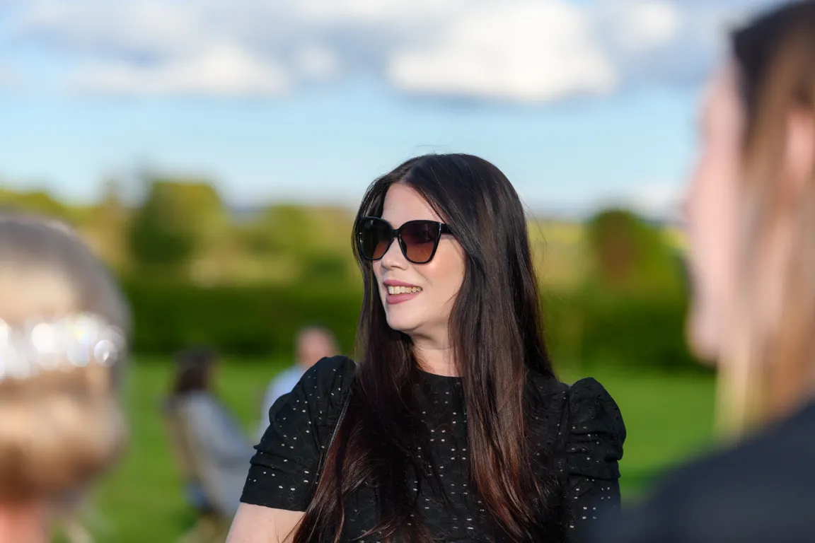 Woman with long dark hair wearing sunglasses and a black eyelet top smiling outdoors with blurred people and greenery in the background.