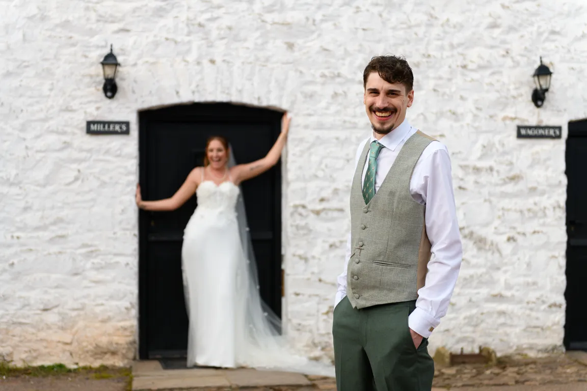 Smiling groom in green tie and vest stands in front of bride in wedding dress leaning on black door with white stone wall background.