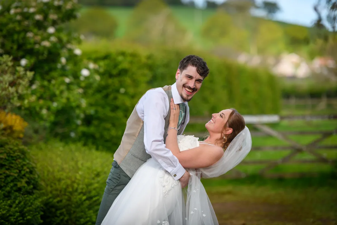 Smiling groom in beige vest and bride in white dress and veil embracing outdoors with greenery and wooden fence in background.