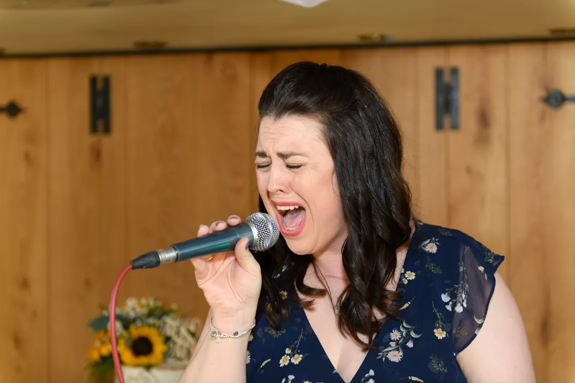 Woman with dark hair passionately singing into a microphone against a wooden wall background.