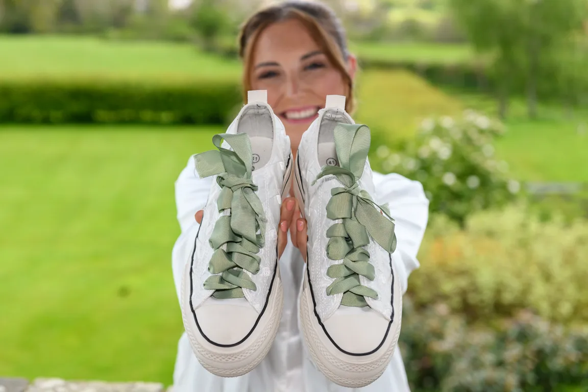 White sneakers with green ribbon laces held up by a smiling woman against a blurred garden background.