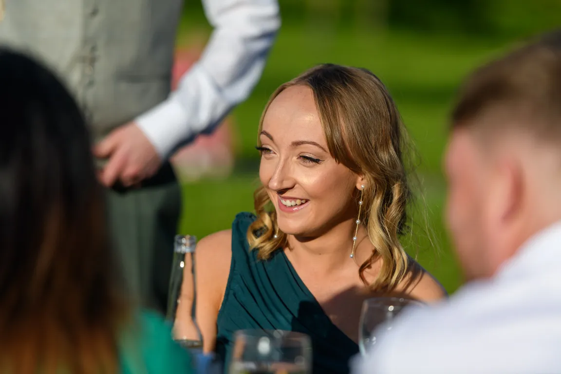 Smiling woman with wavy blonde hair wearing a blue sleeveless dress and dangling pearl earrings at an outdoor gathering.