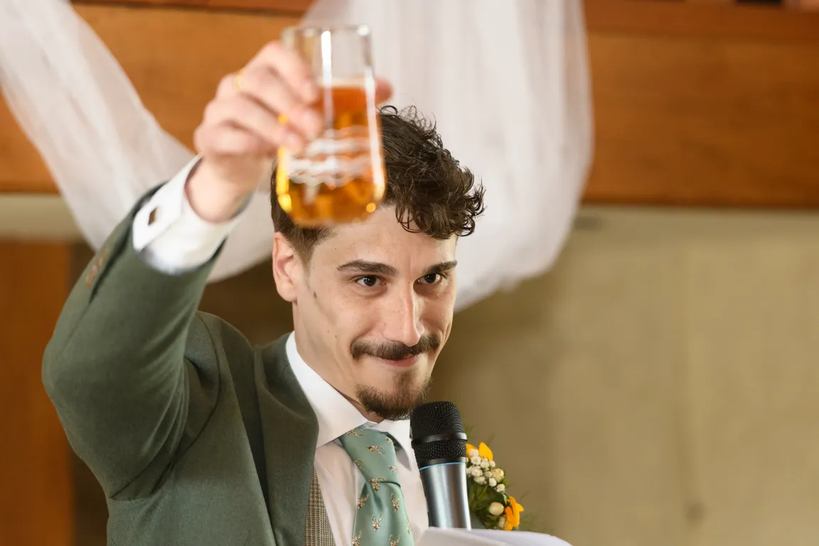 Man in a green suit and teal deer-patterned tie holding up a glass of amber liquid while speaking into a microphone.