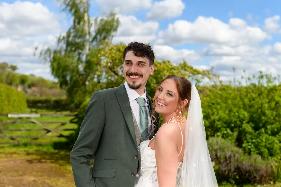 Smiling bride in white dress and veil leaning on groom in green suit and floral tie outdoors with green foliage and blue sky in the background.
